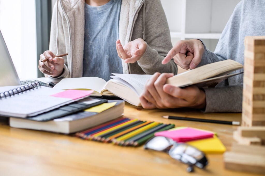 Zwei Personen sitzen an einem Tisch mit aufgeschlagenen Büchern, Stiften und Notizzetteln und diskutieren