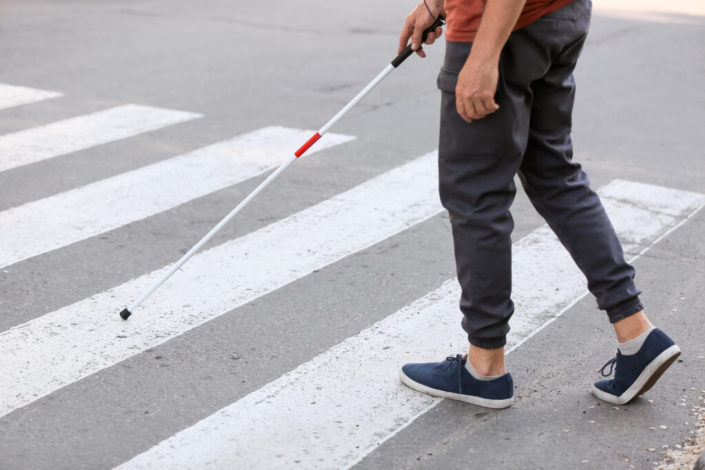 Person mit dunkler Hose und blauen Schuhen tastet mit weißem Blindenstock einen Zebrastreifen ab