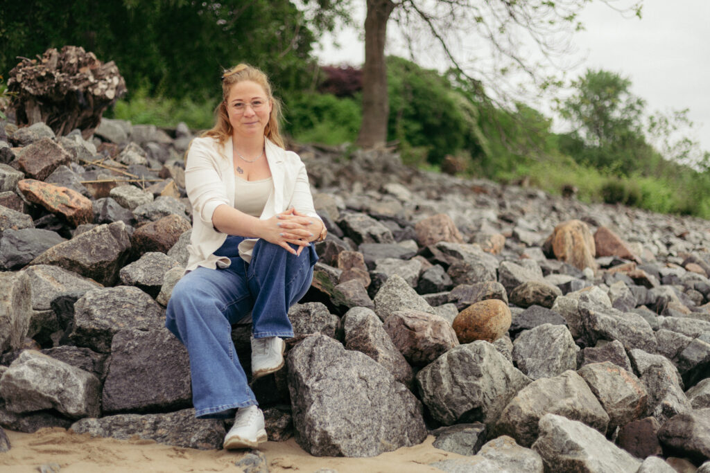 Melanie sitzt auf Steinen am Strand lächelt in die Kamera