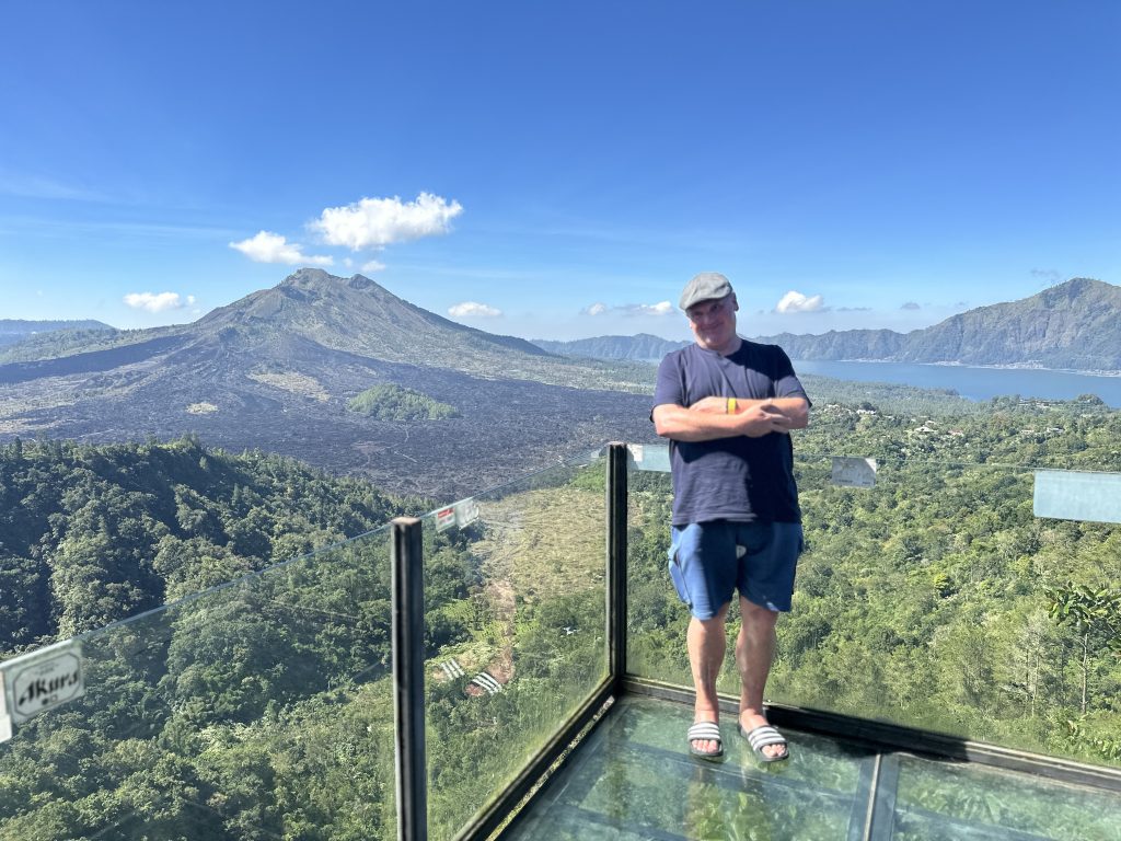 Bernd Leitz steht mit verschränkten Armen auf einem gläsernen Aussichtspunkt mit Blick auf den Mount Batur und den Batur-See im Hintergrund auf Bali